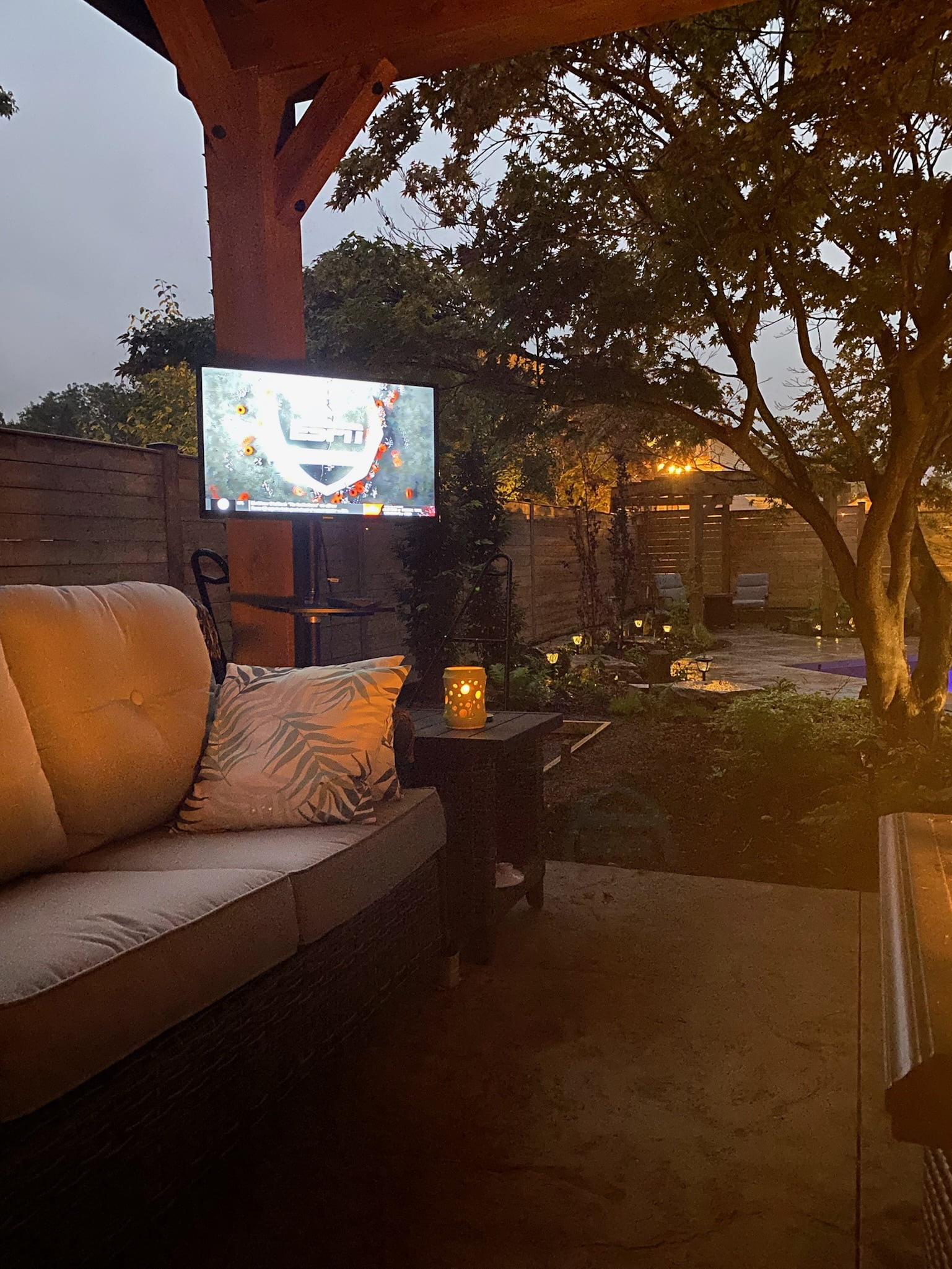 covered outdoor seating area by pool with tv at night