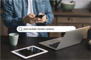 person sitting at table with laptop and tablet holding mobile phone searching pool builder london ontario